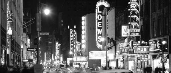 Une rue, la nuit, des voitures et des piétons circulent entre les affiches illuminées des salles de théâtre et de cinéma 