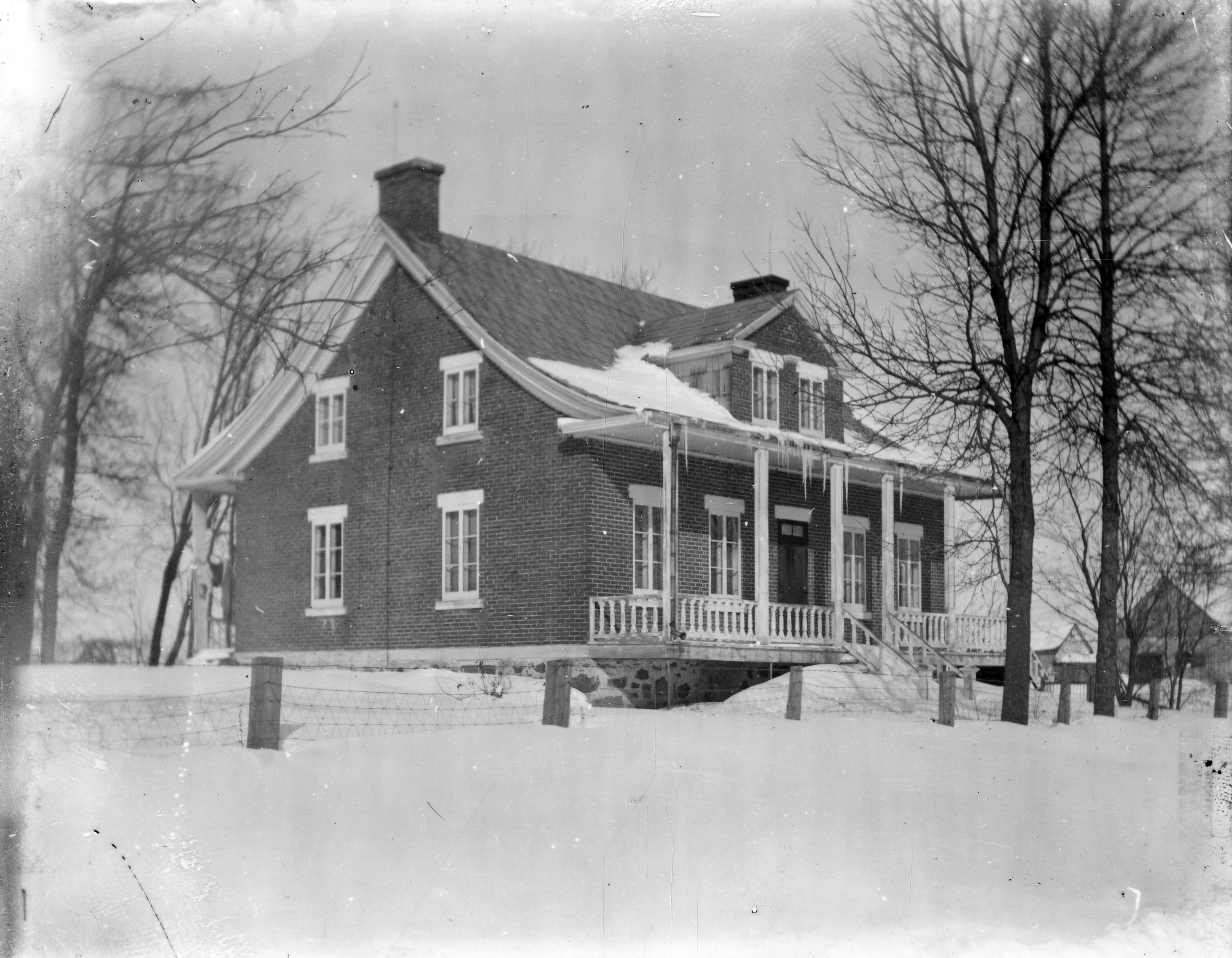 Photographie en noir et blanc. Vue d'une maison l'hiver. Des glaçons sont suspendus au toit et deux arbres dénudés de feuilles ornent l'entrée