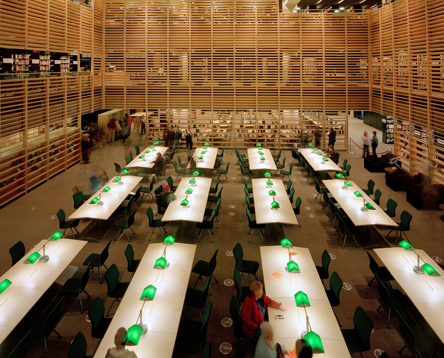 Salle de lecture meublée avec de longues tables et de lampes vertes.