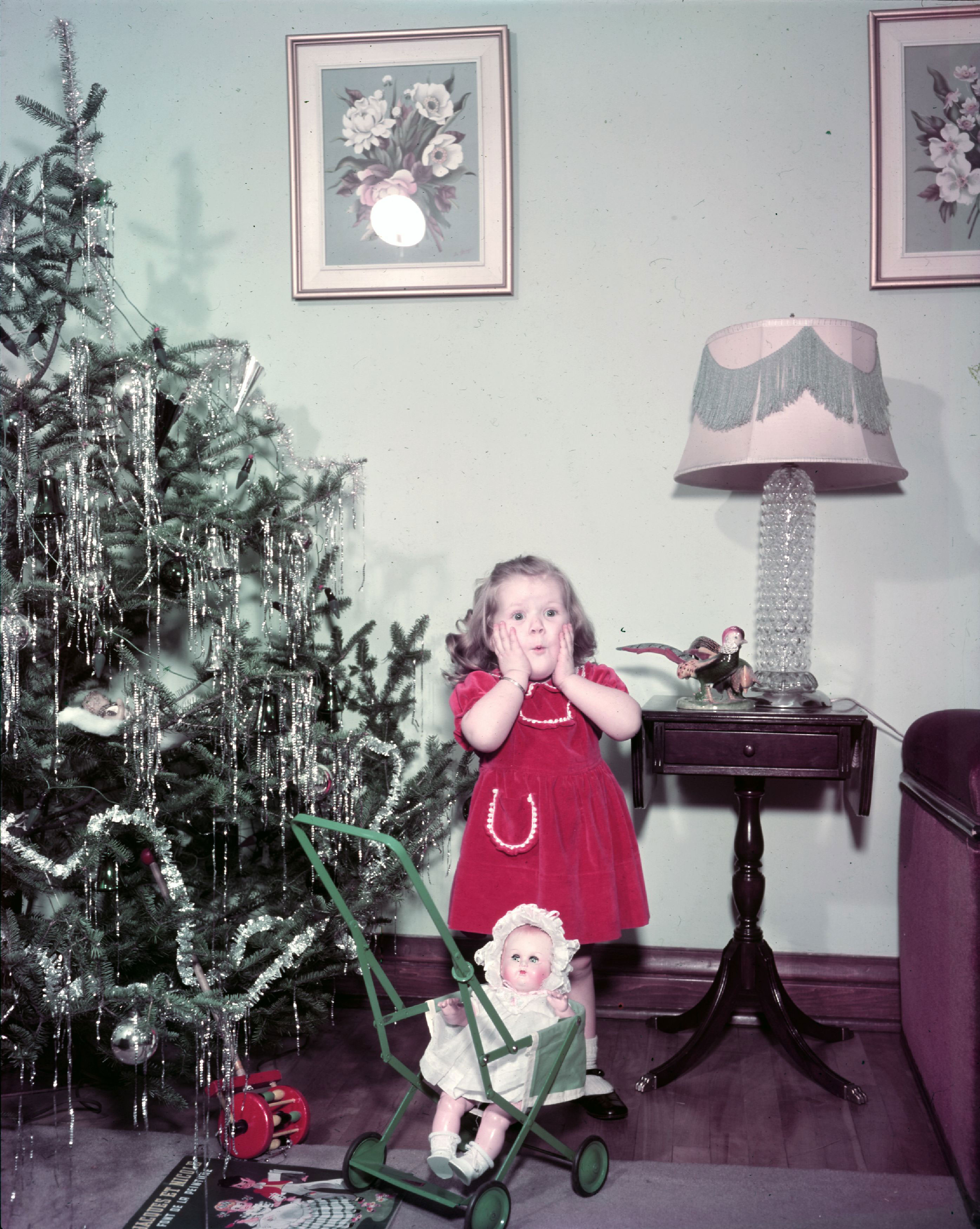 Fillette entourée de jouets, se tenant à côté d'un arbre de Noël.