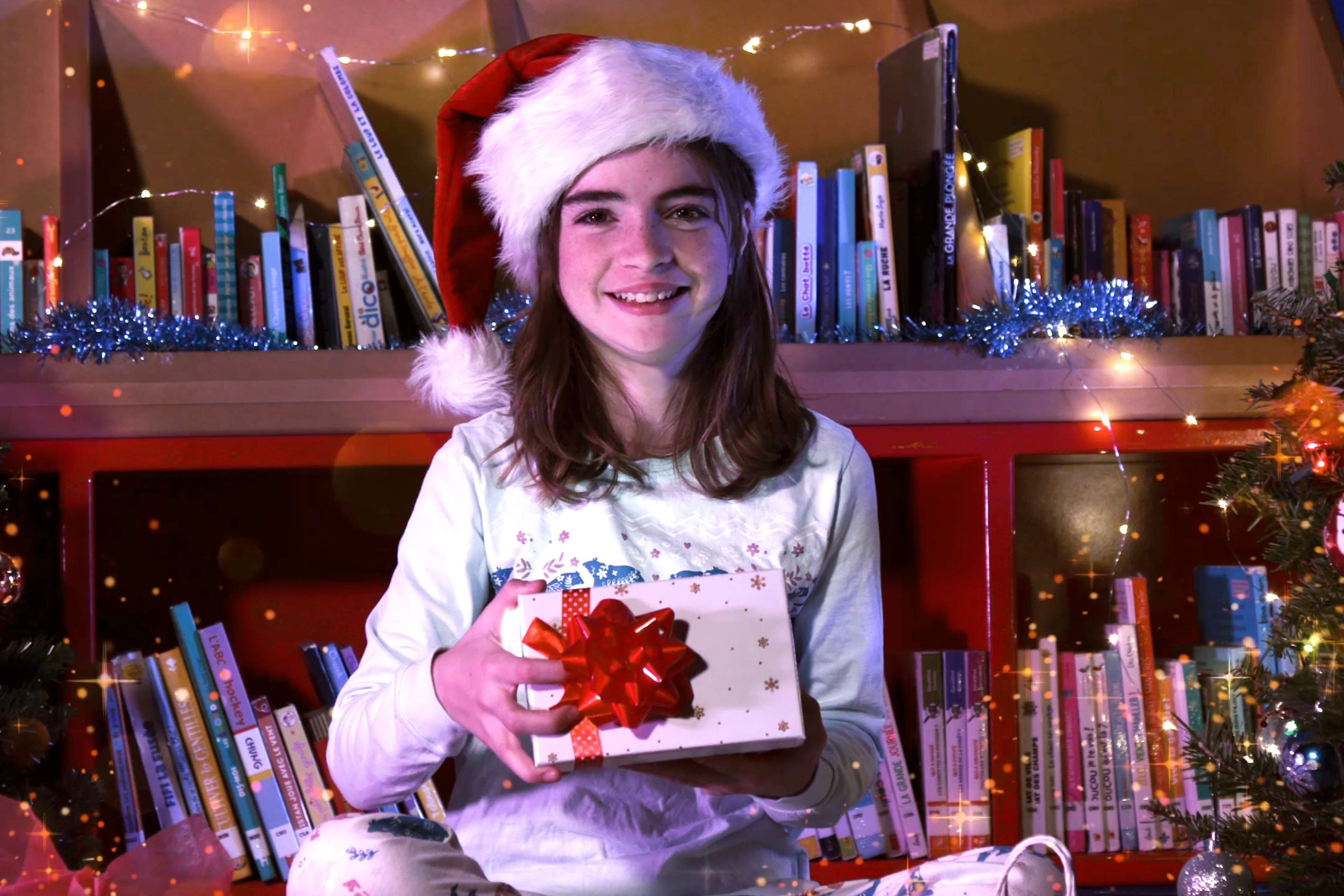 Petite fille avec un chapeau de Noël qui sourit en ouvrant une boîte cadeau entourée de livres et de cadeaux au pied du sapin.