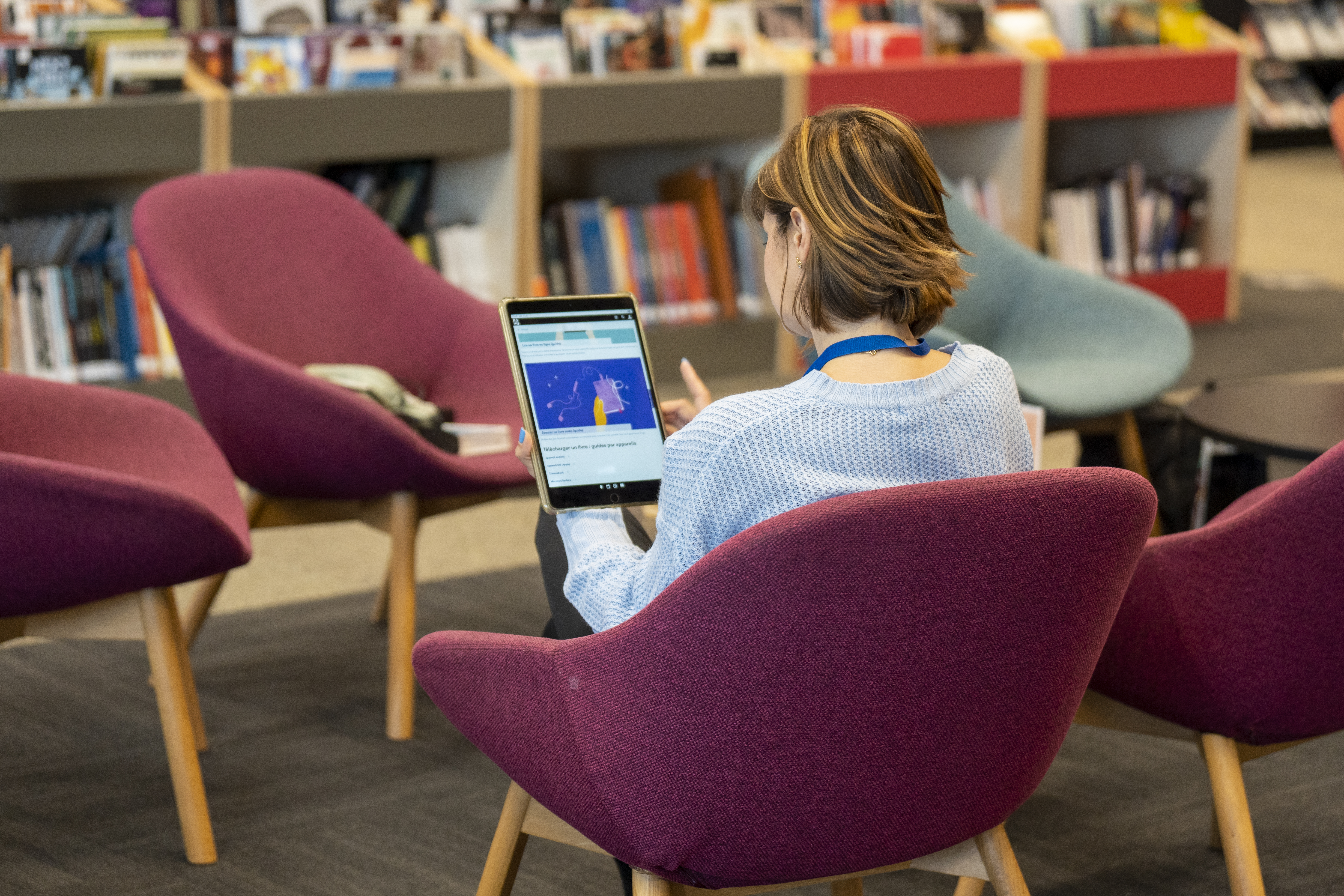 Femme de dos, assise dans un fauteuil, une tablette numérique dans les mains.