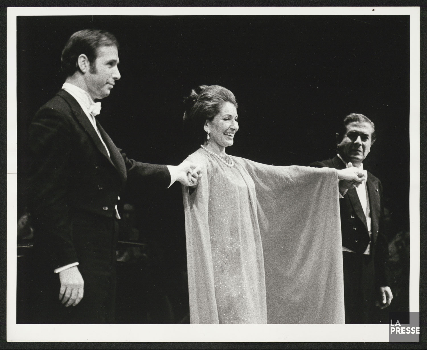 Une femme en robe de soirée, souriant, entourée de deux hommes en smoking lui tenant la main.