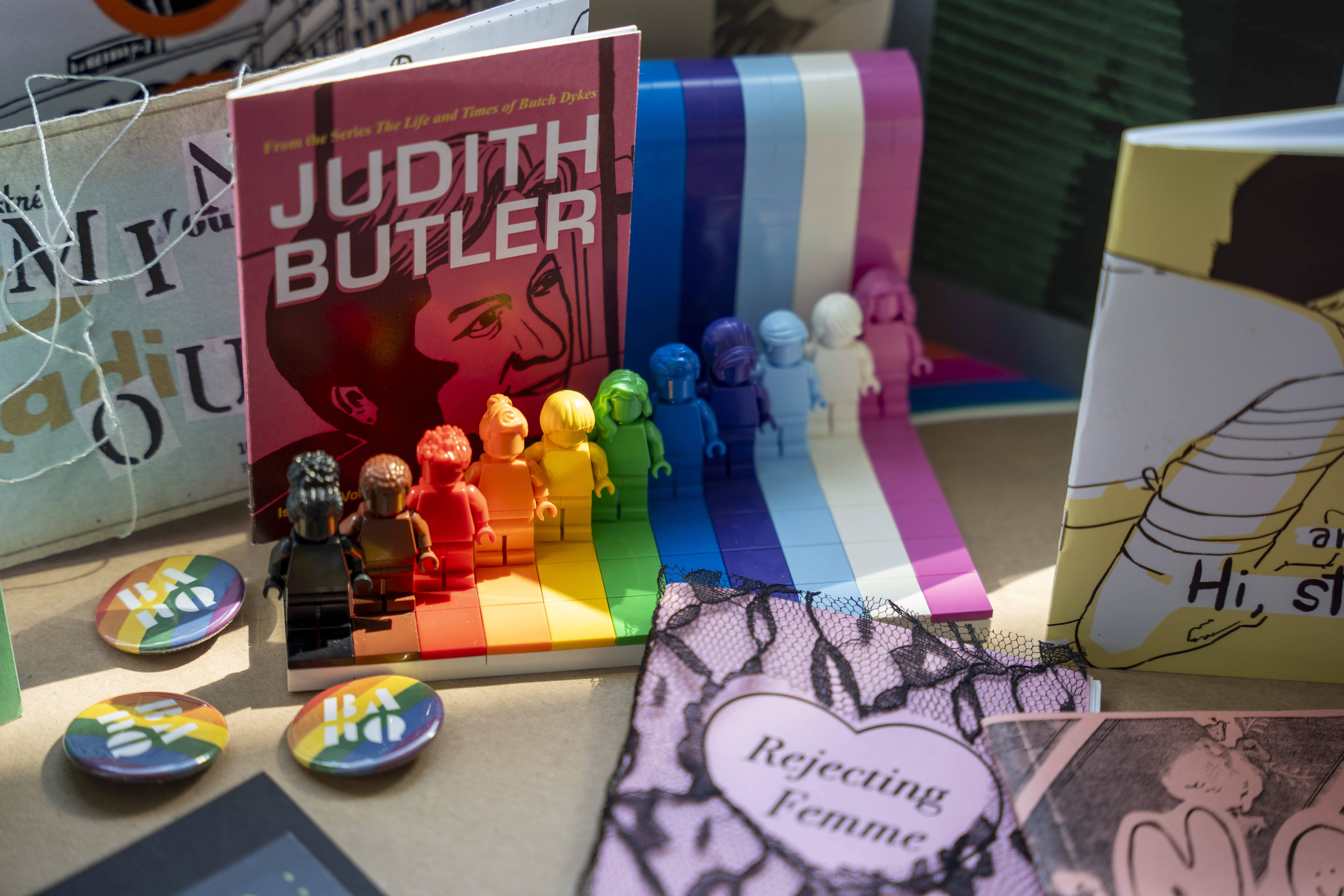 Quelques zines dont « Judith Butler » et » Rejecting Femme », macarons de BAnQ aux couleurs de l'arc-en-ciel et personnages Lego de différentes couleurs.