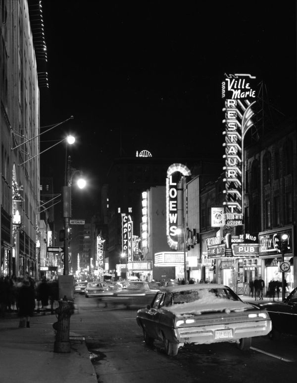 Une rue, la nuit, des voitures et des piétons circulent entre les affiches illuminées des salles de théâtre et de cinéma 