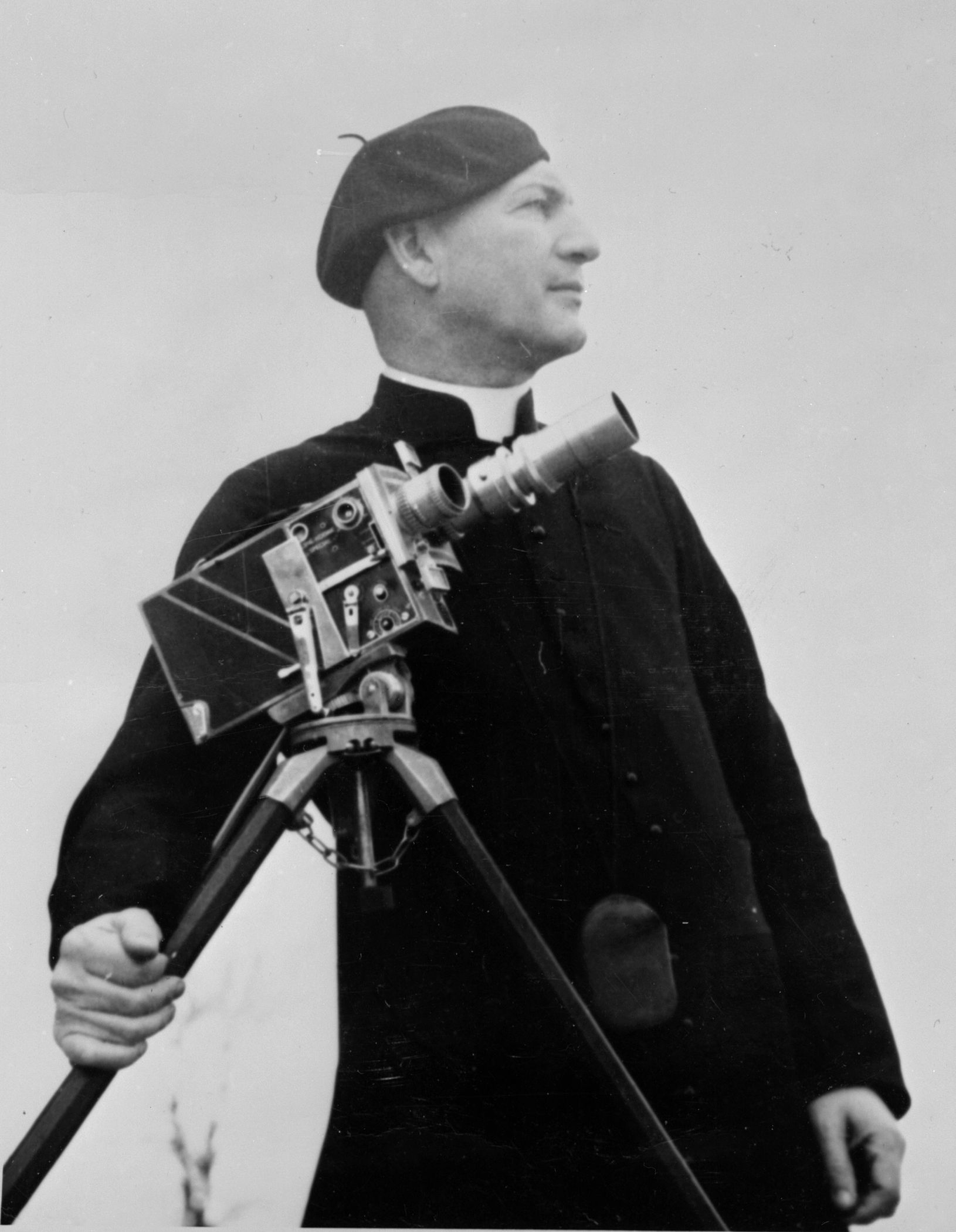 Maurice Proulx portant une soutane et un béret et tenant une caméra.
