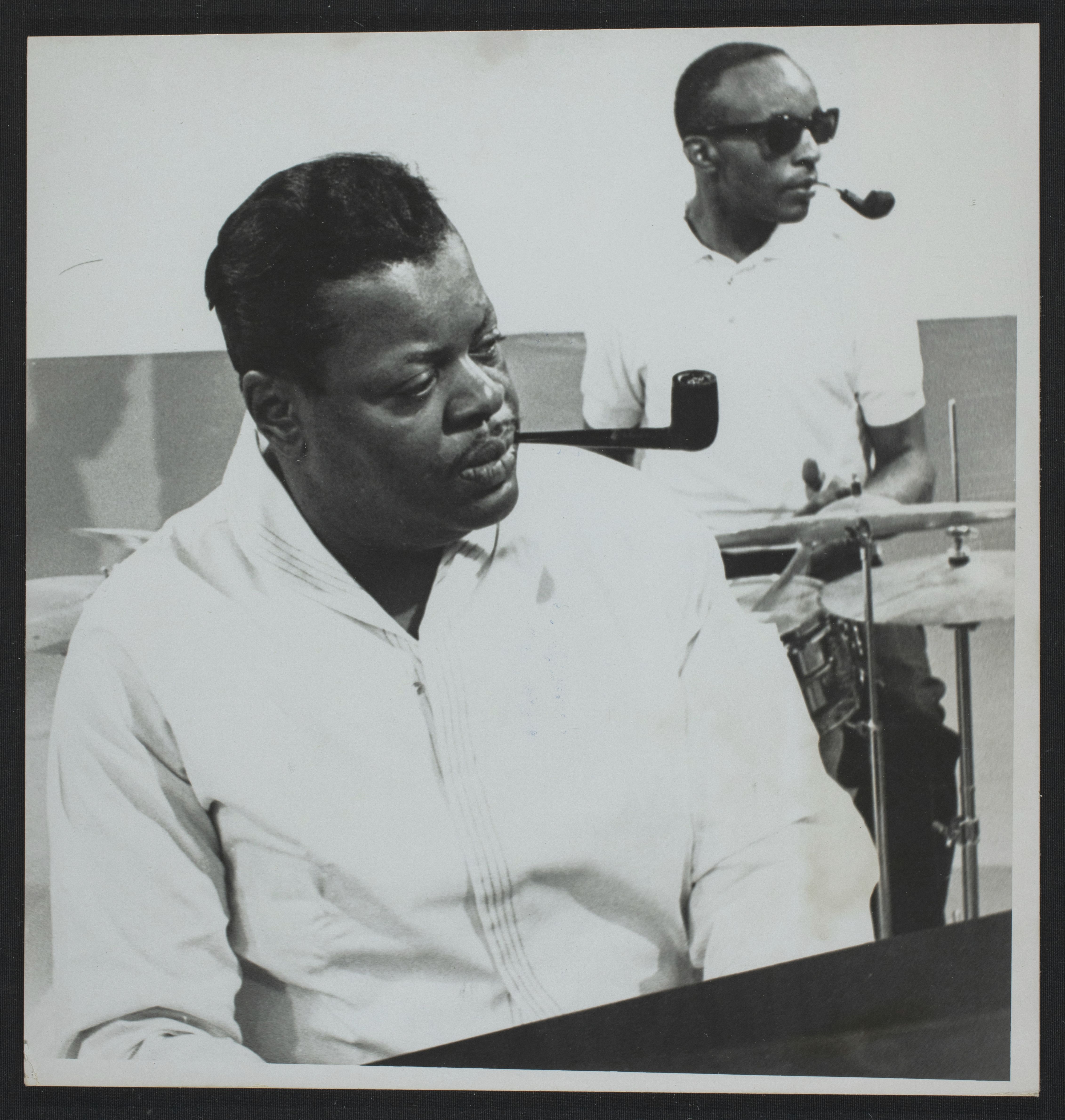 Oscar Peterson, pipe au bec, jouant du piano. Un batteur est visible en arrière-plan. Il porte des lunettes fumées.