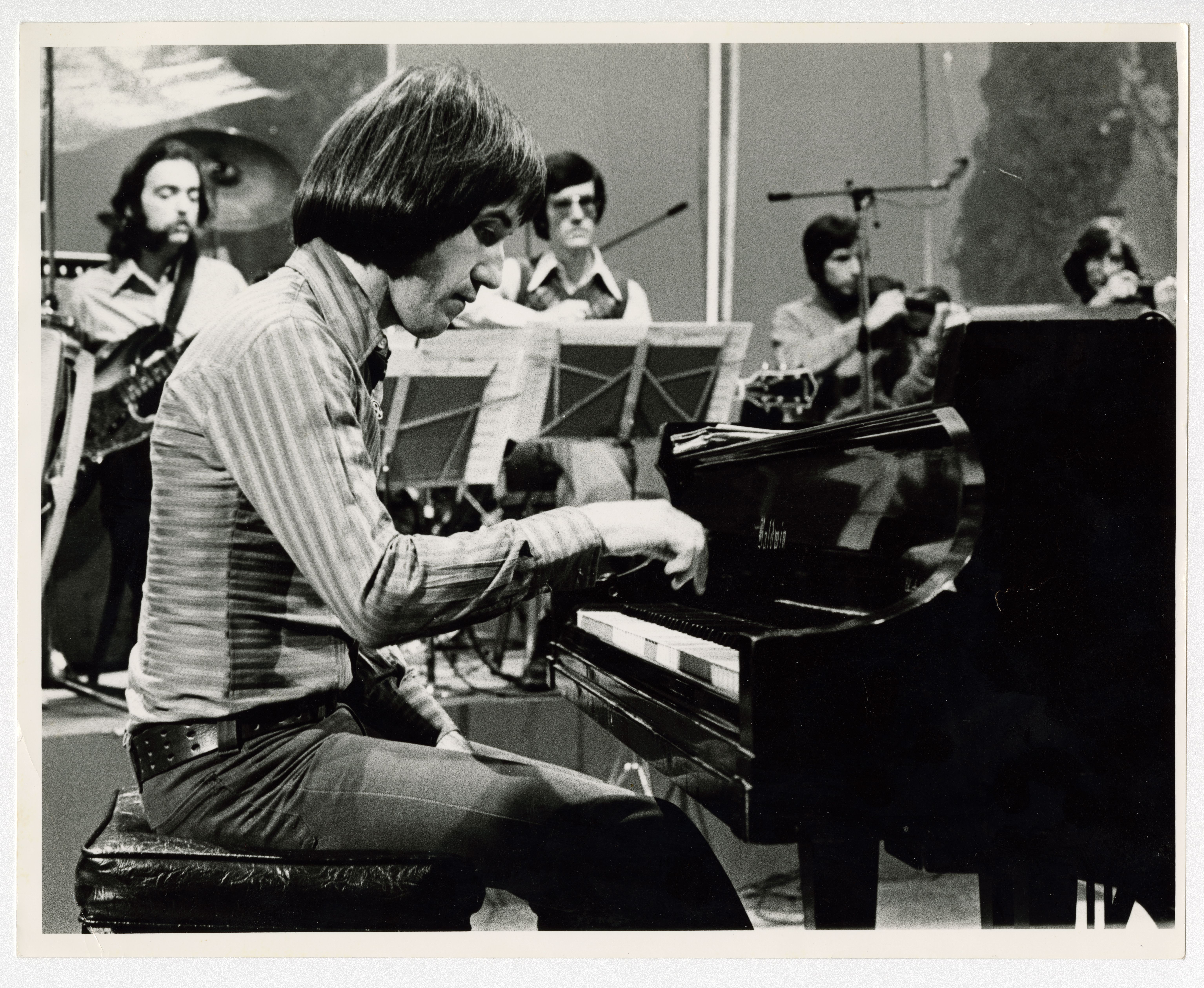 Homme assis à un piano, entouré d'un groupe de musiciens.