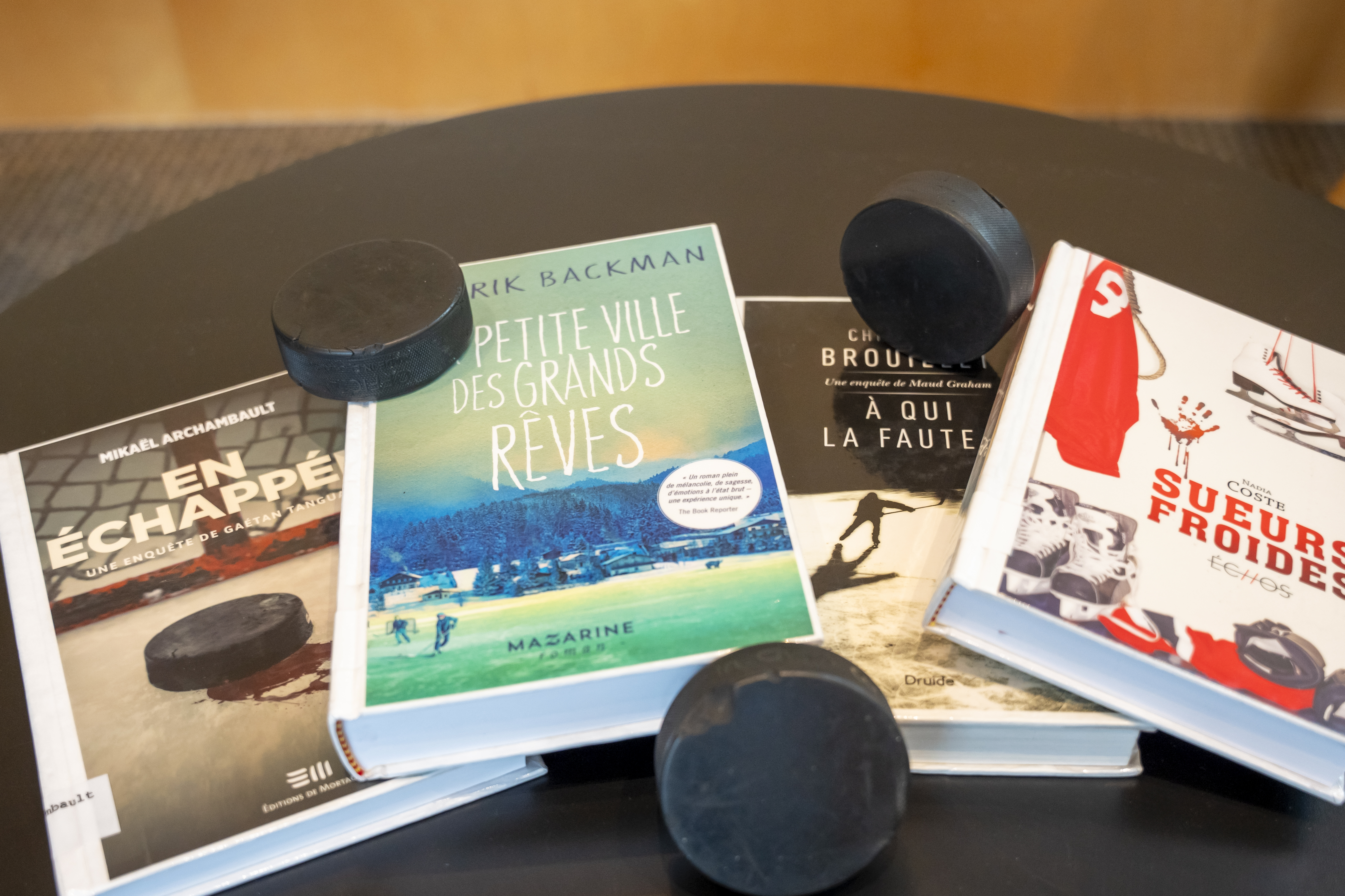 Quatre livres dont l'action tourne autour du hockey sont posés sur une table avec trois rondelles de hockey.
