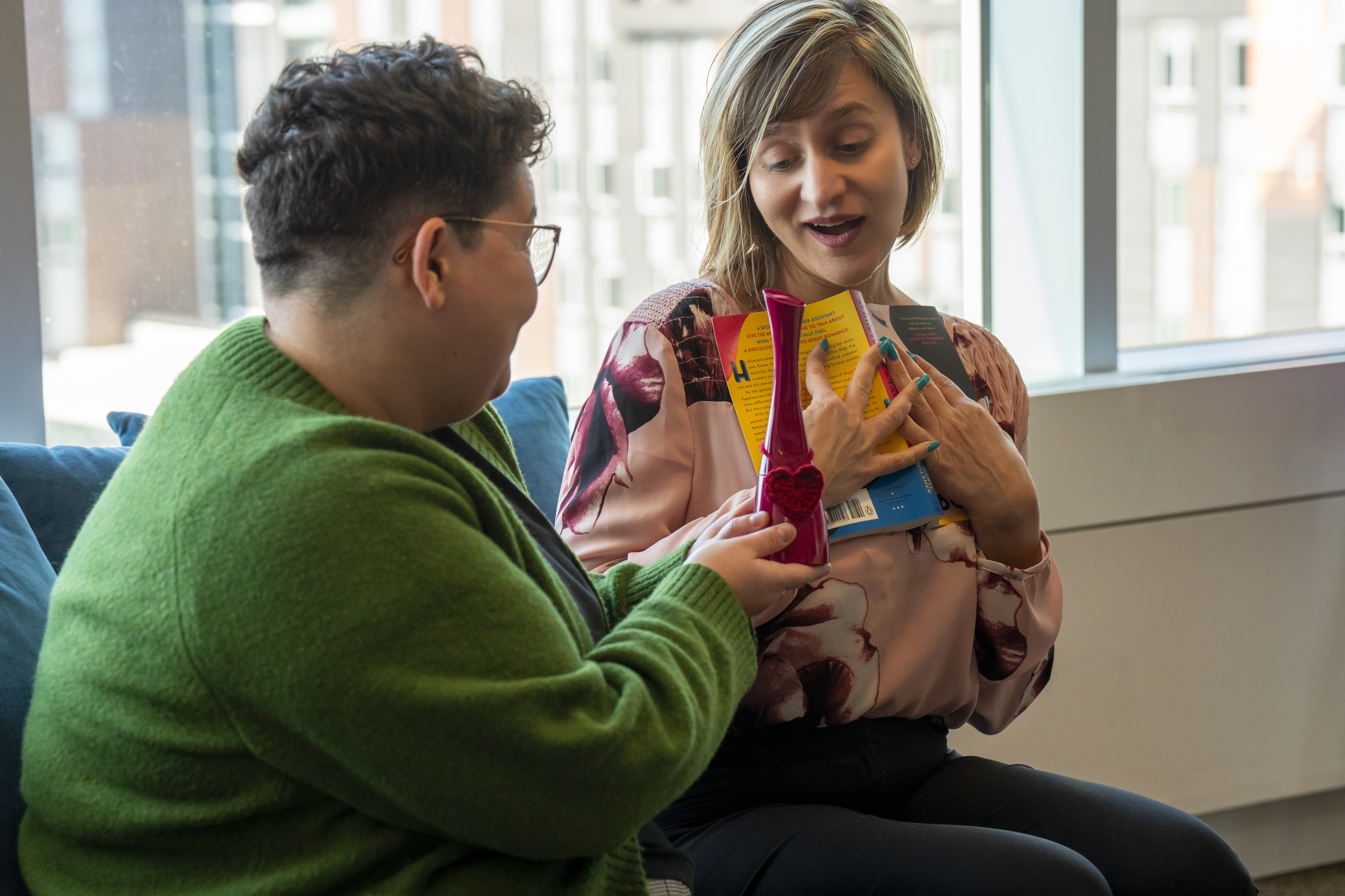 Une femme tend une bouteille de parfum à une autre qui tient un livre.