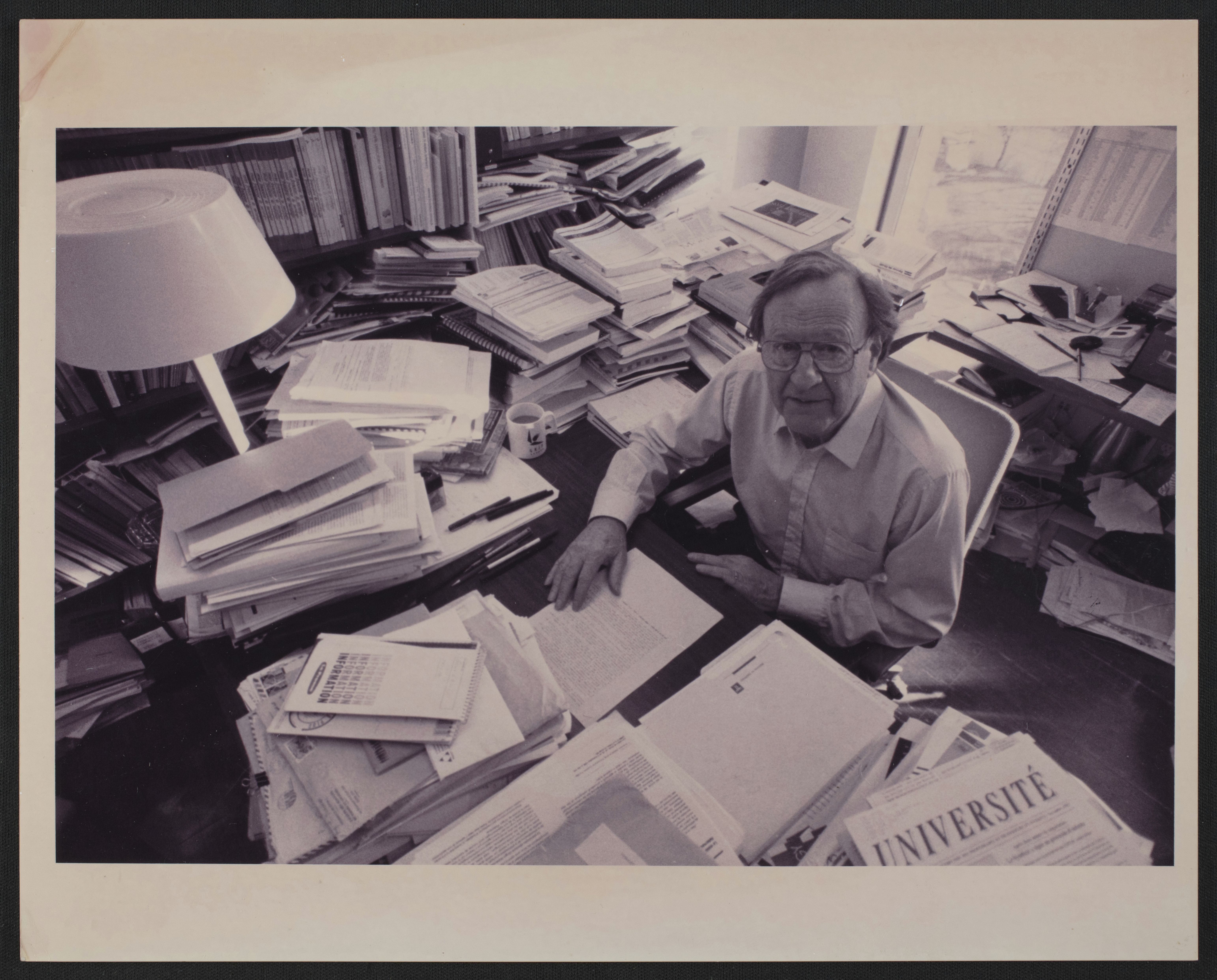Un homme assis à son bureau, entouré de quantité de dossier papier.