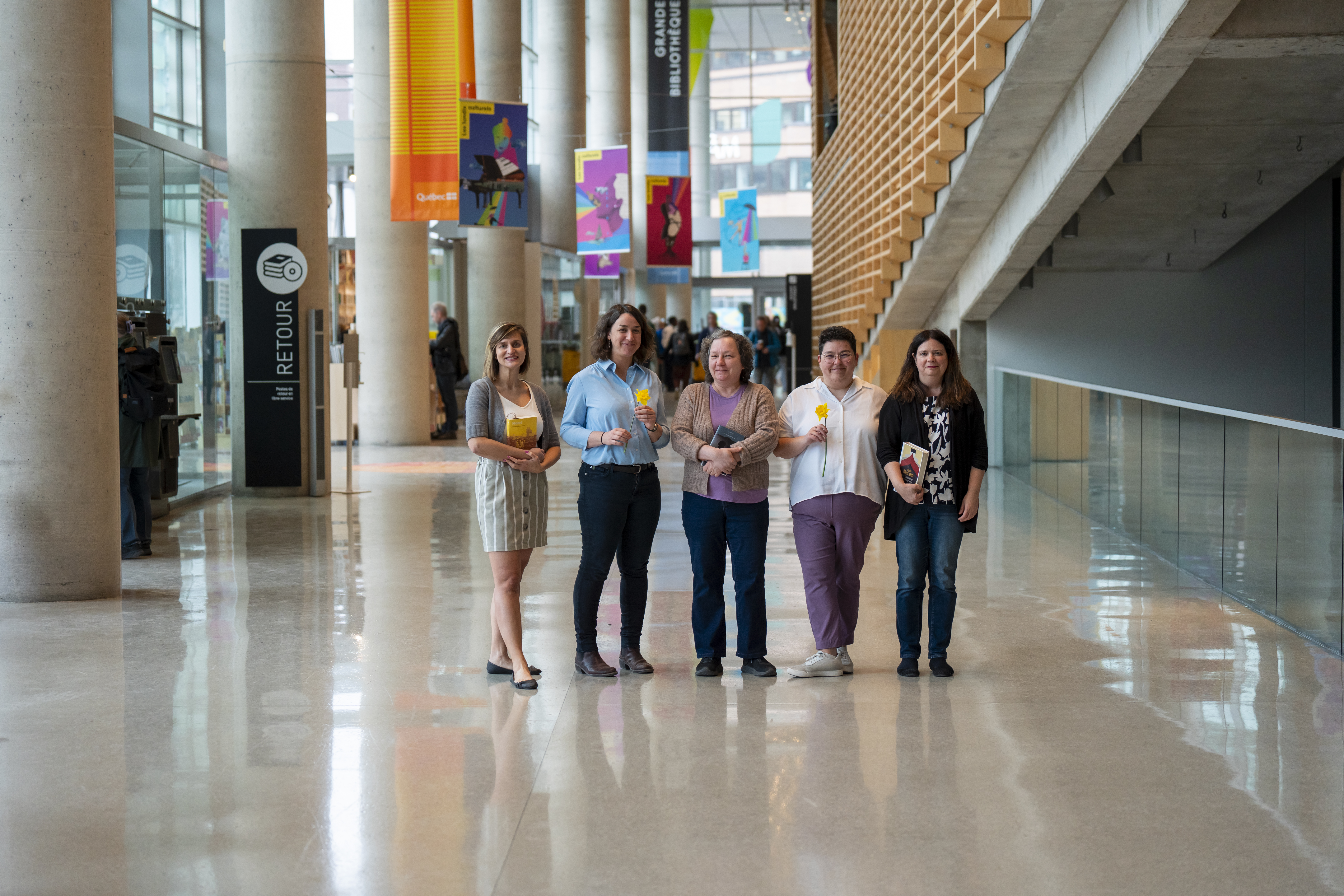 Cinq femmes, debout dans le hall de la Grande Bibliothèque, tiennent des livres dans leurs mains.