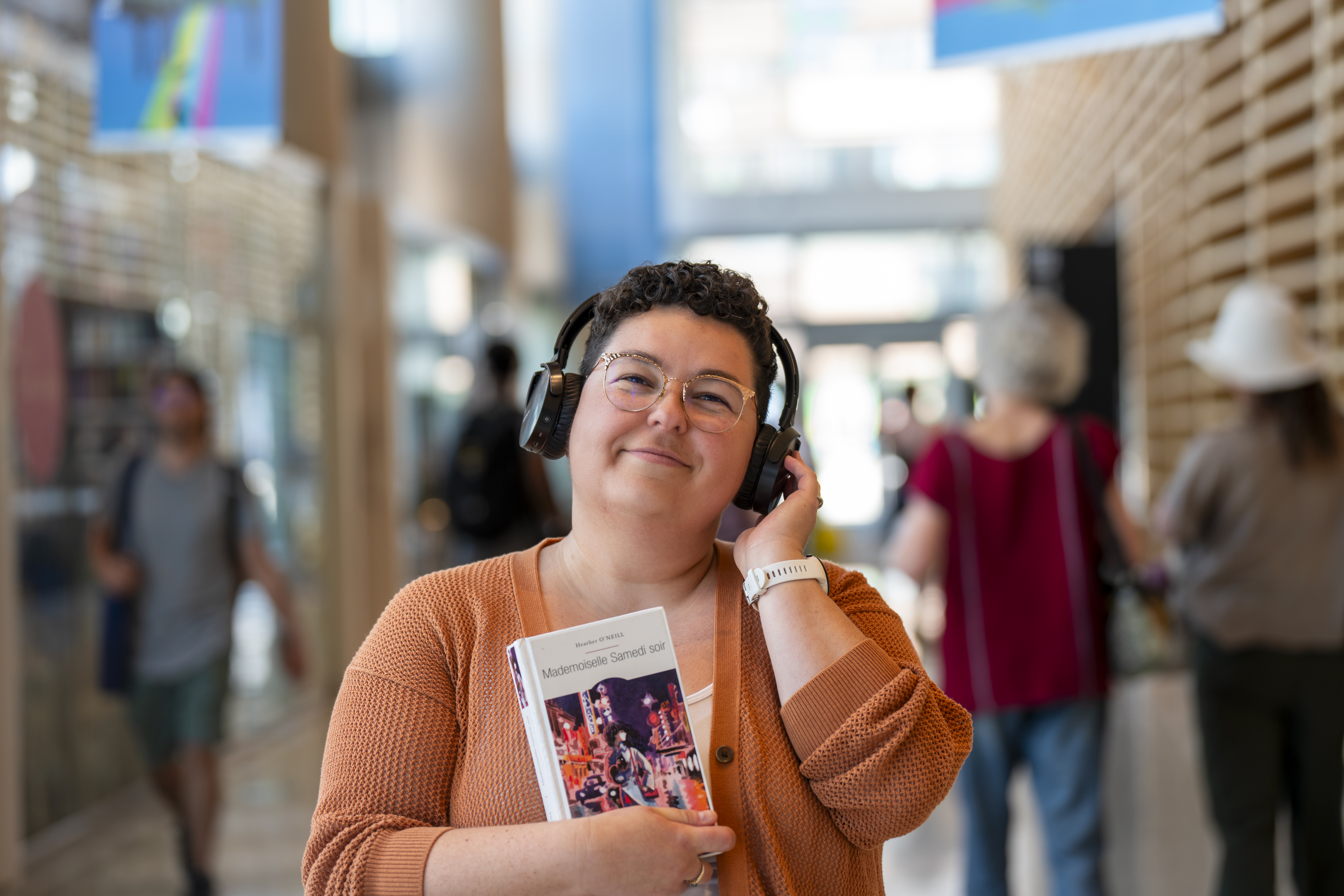 Une femme portant un casque d'écoute tient le livre Mademoiselle Samedi soir, de Heather O’Neill. Elle est dans le hall de la Grande Bibliothèque.