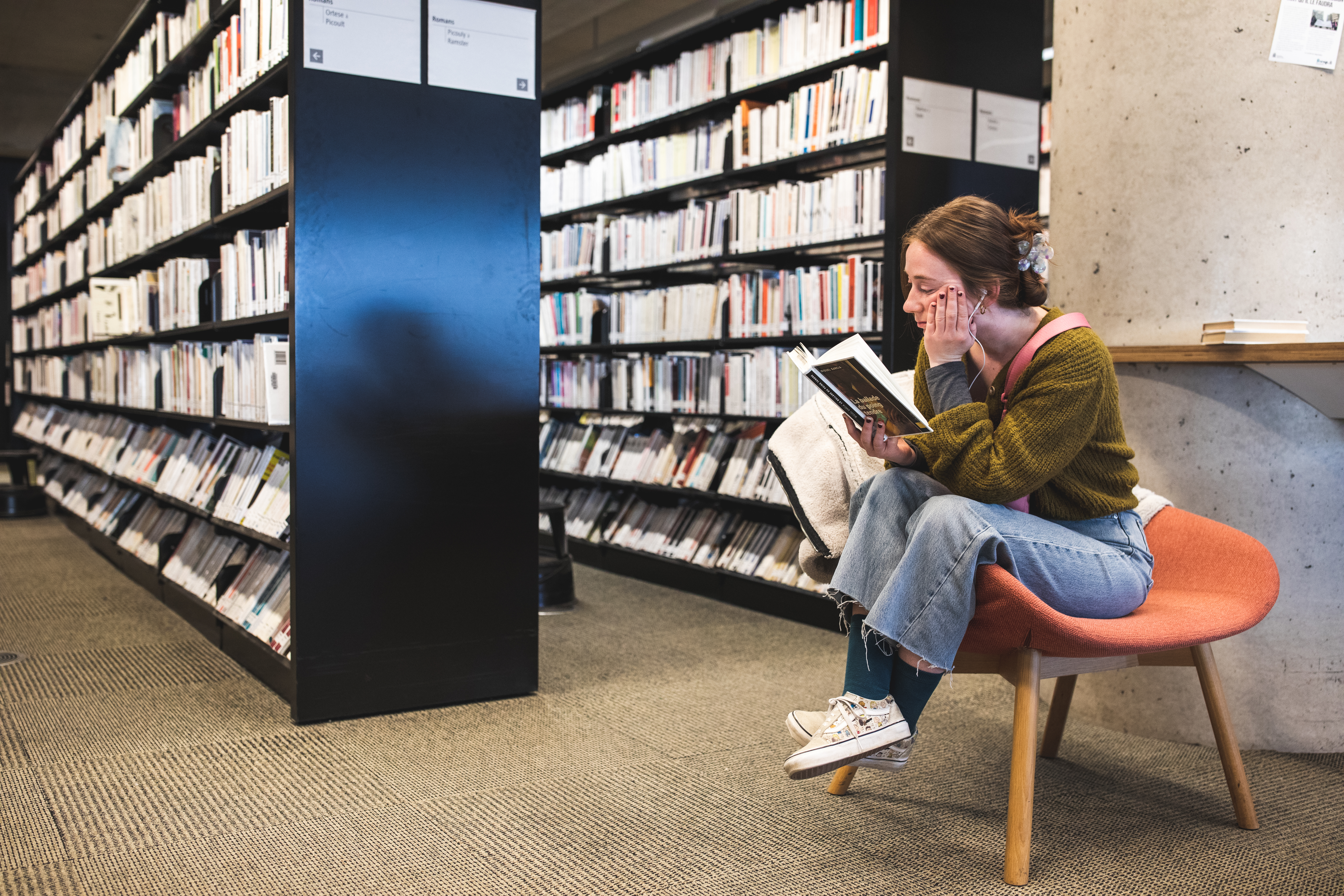 Jeune fille lisant un livre dans un fauteuil près de rayonnages de bibliothèque