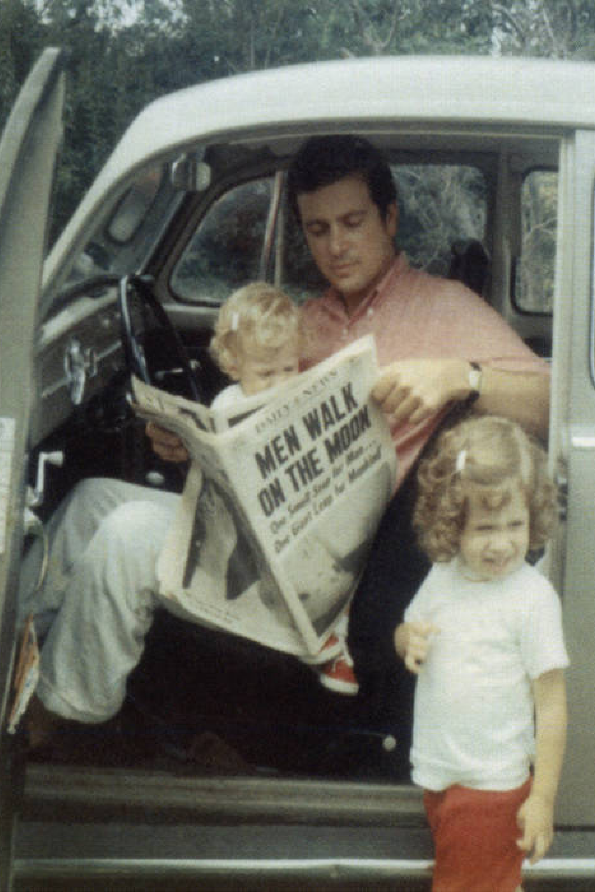 Un père assis dans sa voiture coccinelle lit son journal la portière ouverte avec un enfant sur les genoux et l’autre à côté de la voiture.