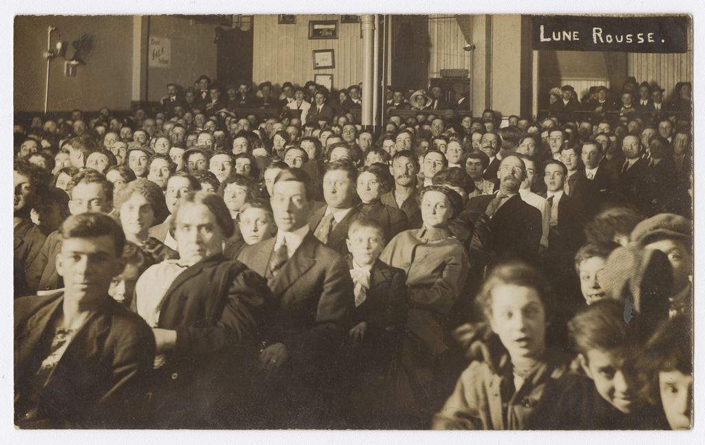 Des dizaines de personnes assises dans une salle accueillant des spectacles et des projections. Les spectateurs fixent l’objectif. 