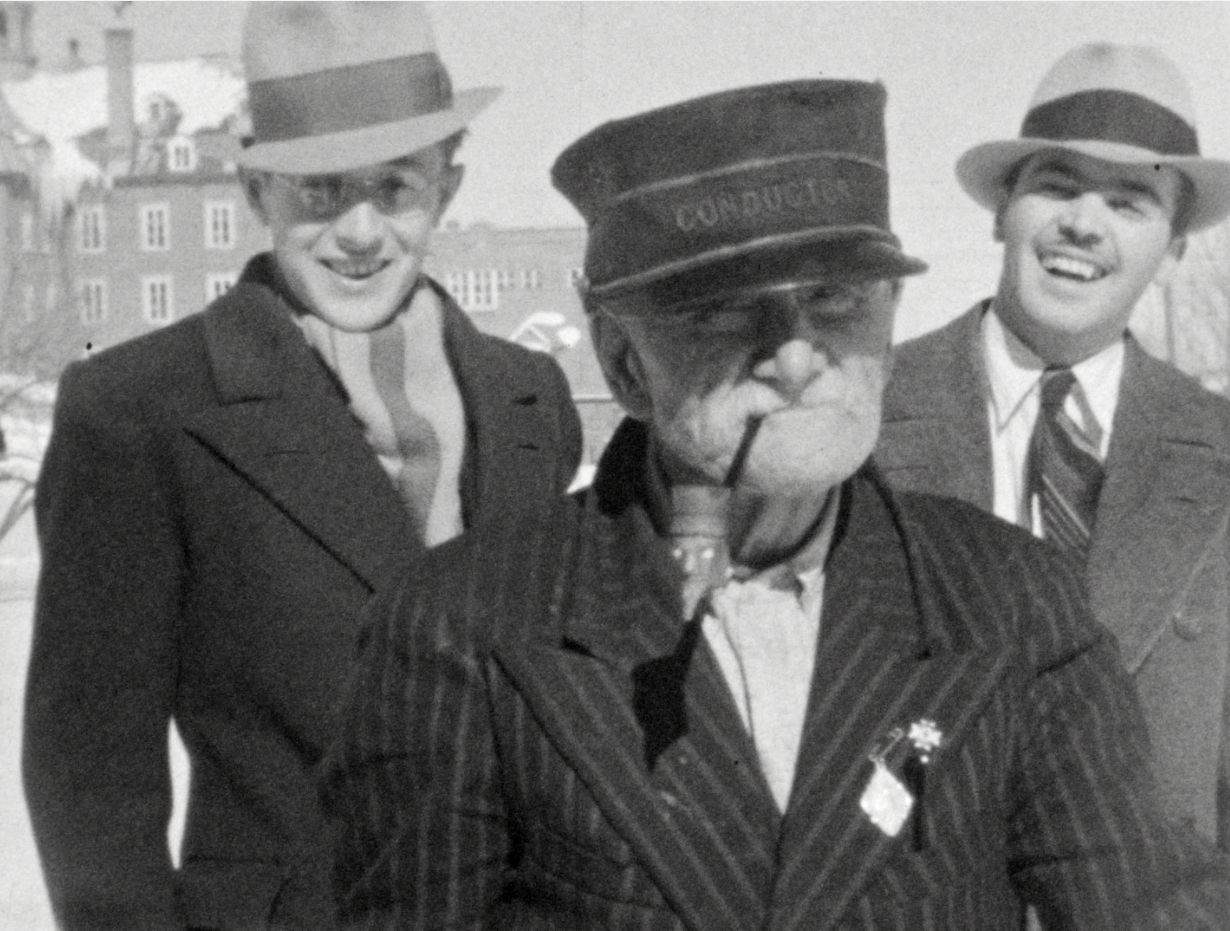 Trois personnes en manteaux et chapeaux posent à l’extérieur devant un bâtiment en arrière-plan.