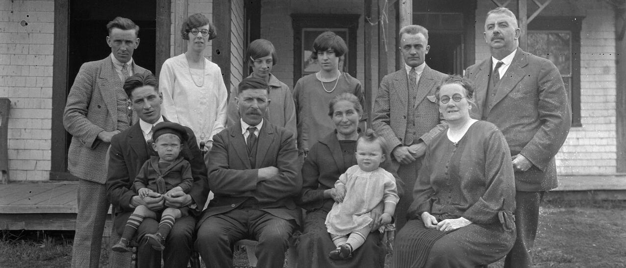 Photographie en noir et blanc d'une famille de 12 personnes placées sur deux rangées. Quatre personnes sont assises. Deux d'entre elles sont sur leur cuisses un bambin. Les six autres personnes sont debout derrière les chaises. 