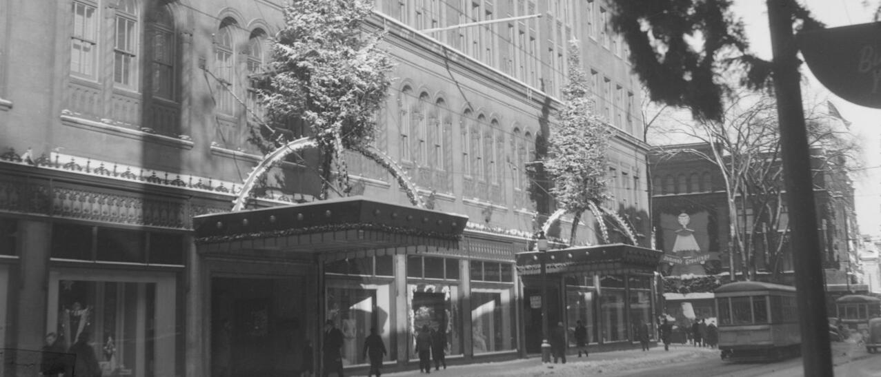 Nous voyons la façade du magasin à rayons Eaton's de la rue Sainte-Catherine Ouest à Montréal. Il y a un arbre de Noël sur les deux marquises des portes d'entrée.