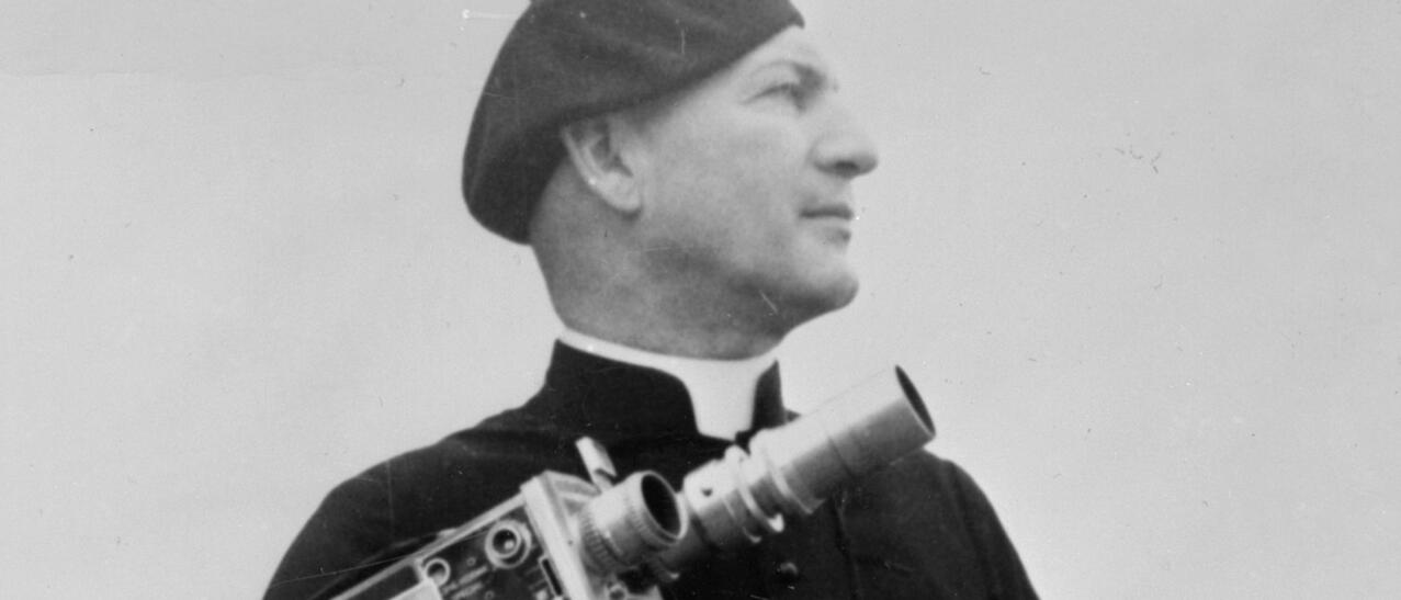 Maurice Proulx portant une soutane et un béret et tenant une caméra.