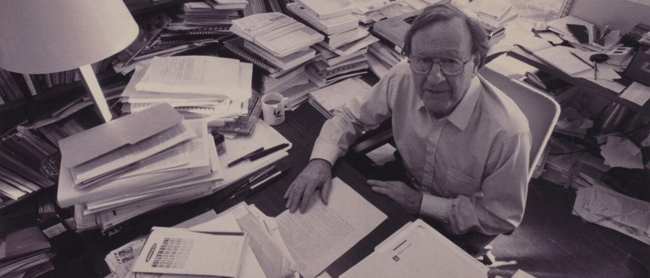 Un homme assis à son bureau, entouré de quantité de dossier papier.