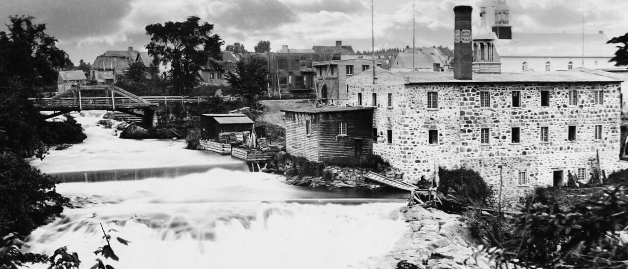 Photo noir et blanc montrant un édifice en briques pâles près d'une rivière.e