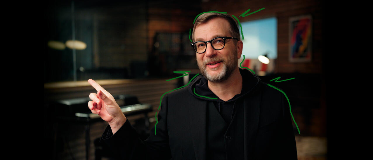 Luc De Larochellière en studio souriant et pointant son doigt vers la gauche