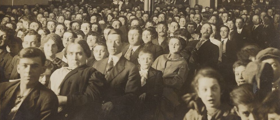 Des dizaines de personnes assises dans une salle accueillant des spectacles et des projections. Les spectateurs fixent l’objectif. 