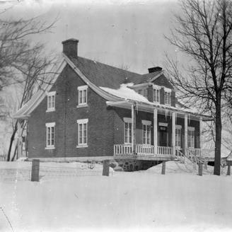 Photographie en noir et blanc. Vue d'une maison l'hiver. Des glaçons sont suspendus au toit et deux arbres dénudés de feuilles ornent l'entrée