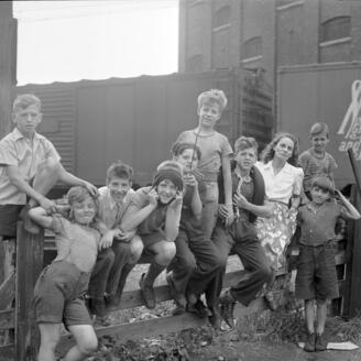 Gabrielle Roy entourée d'enfants dans le quartier Saint-Henri à Montréal. En arrière-plan des wagons de marchandise