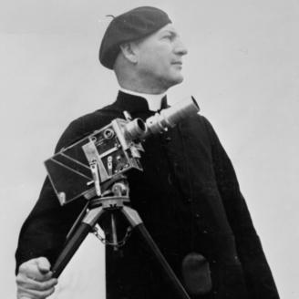 Maurice Proulx portant une soutane et un béret et tenant une caméra.