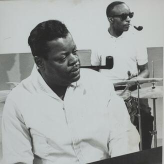 Oscar Peterson, pipe au bec, jouant du piano. Un batteur est visible en arrière-plan. Il porte des lunettes fumées.