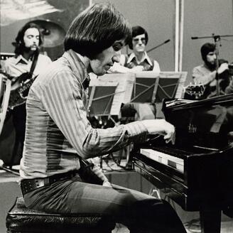 Homme assis à un piano, entouré d'un groupe de musiciens.