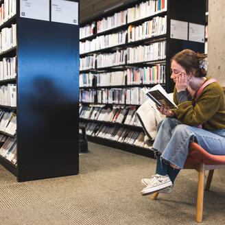 Jeune fille lisant un livre dans un fauteuil près de rayonnages de bibliothèque