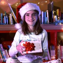 Petite fille avec un chapeau de Noël qui sourit en ouvrant une boîte cadeau entourée de livres et de cadeaux au pied du sapin.