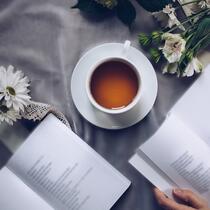 Une personne dont on ne voit que les mains déposées sur les pages est en train de lire un livre qui repose près d'un autre livre ouvert, d'une tasse de thé et de marguerites