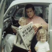 Un père assis dans sa voiture coccinelle lit son journal la portière ouverte avec un enfant sur les genoux et l’autre à côté de la voiture.