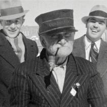 Trois personnes en manteaux et chapeaux posent à l’extérieur devant un bâtiment en arrière-plan.