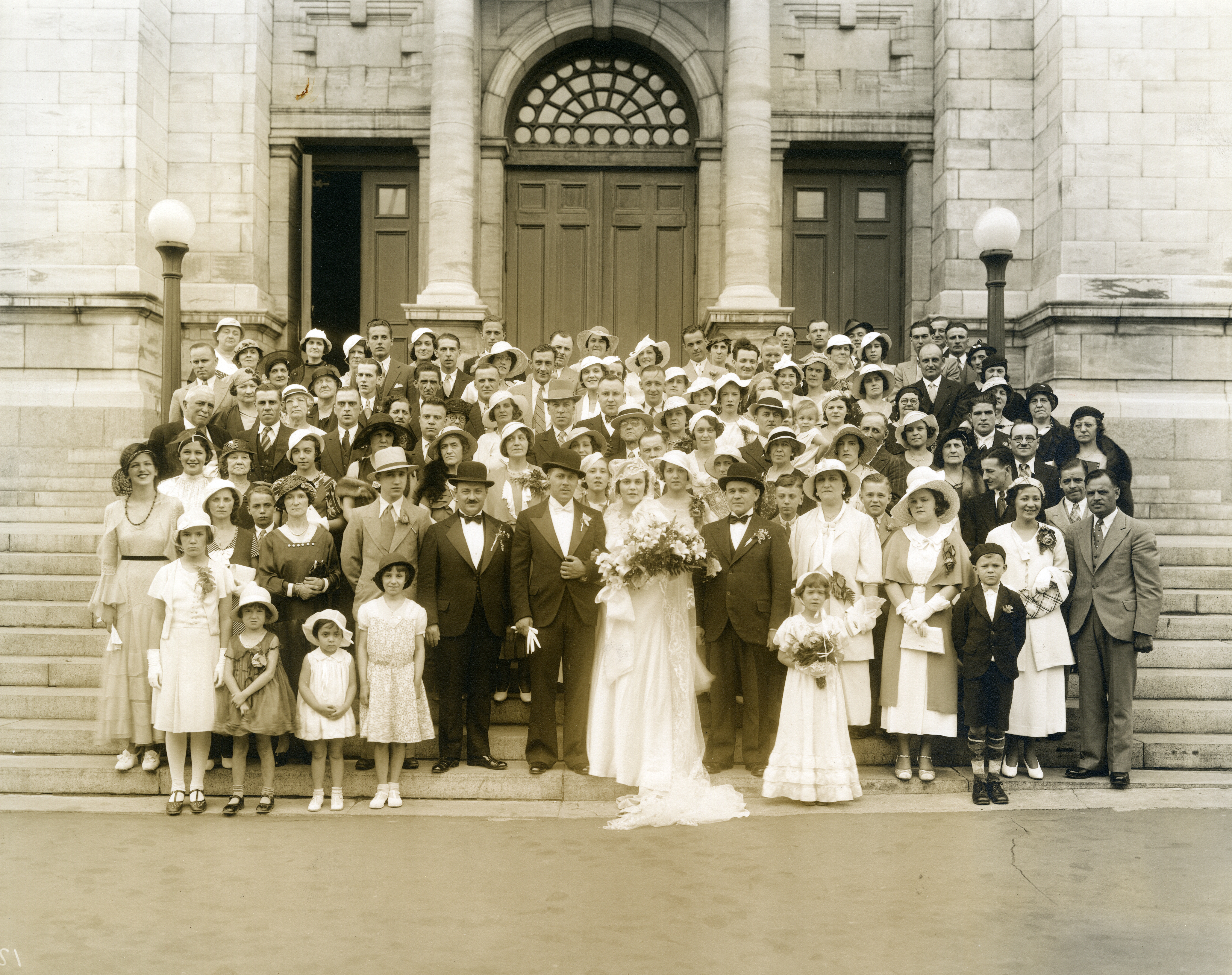 Photographie d'un couple de mariés et de leur famille se tenant devant la façade église. 