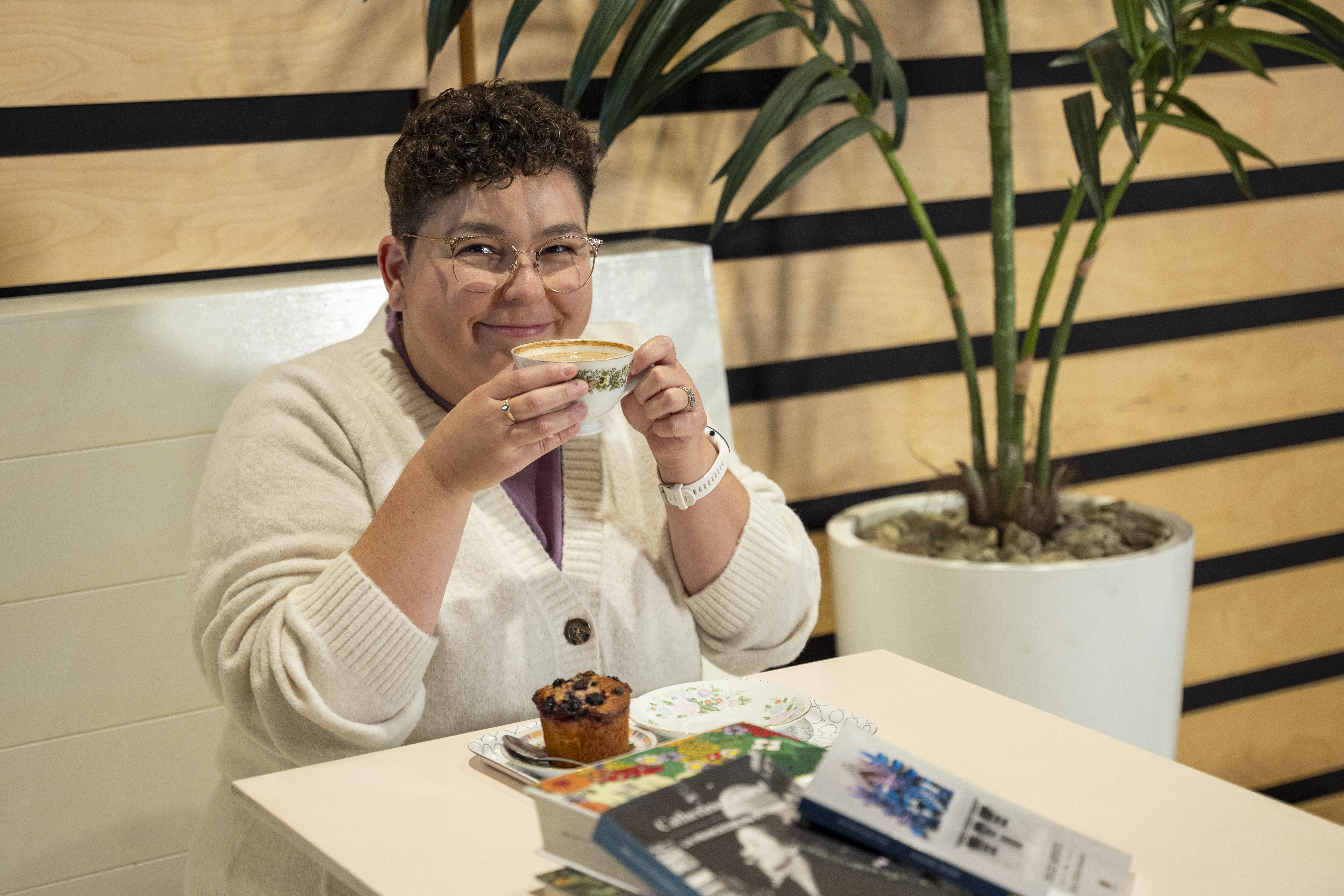 Une femme boit un café, attablée devant des livres.