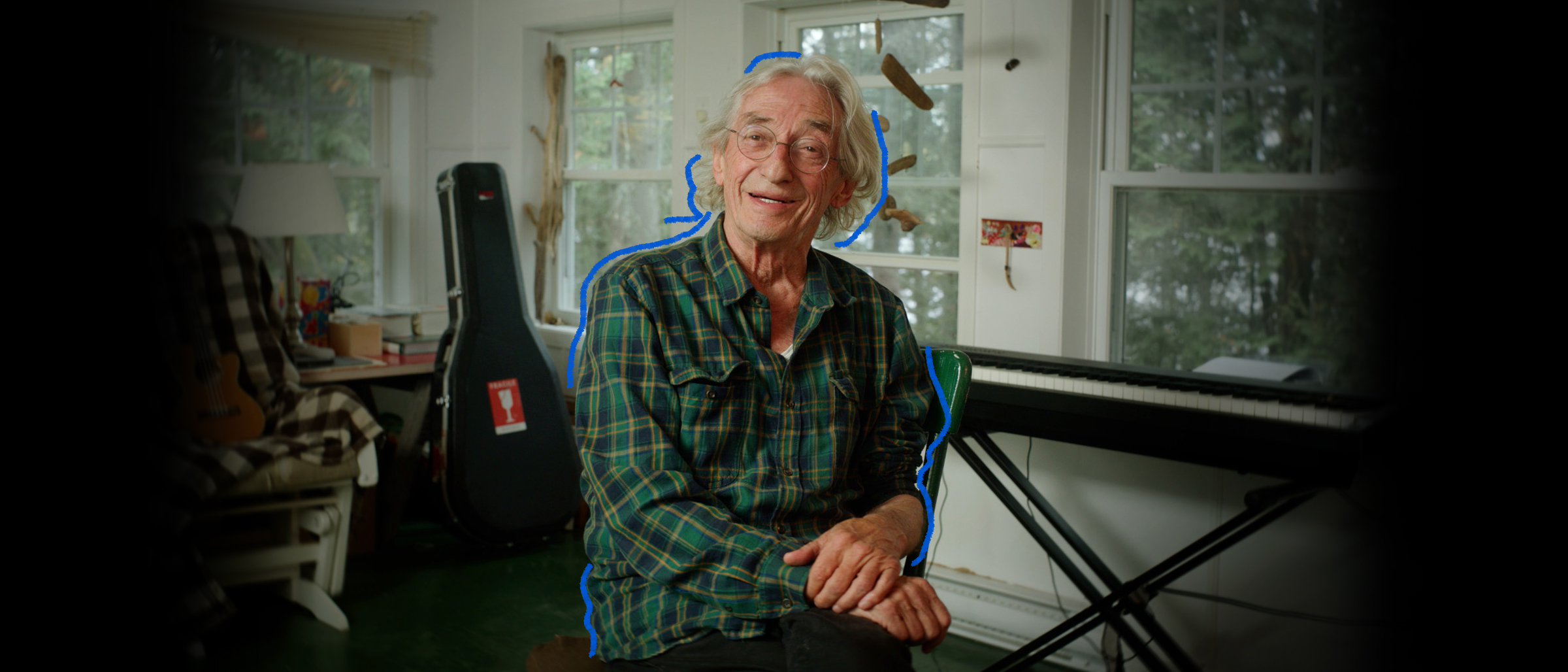 Le chanteur Richard Desjardins, souriant, entouré d'un piano et d'une guitare dans un chalet