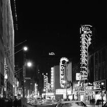 Une rue, la nuit, des voitures et des piétons circulent entre les affiches illuminées des salles de théâtre et de cinéma 